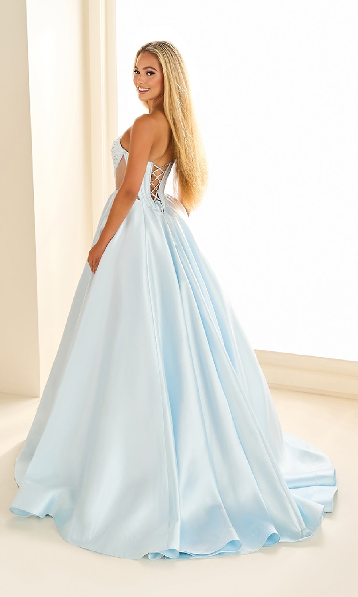 A woman wearing the Ellie Wilde EW36025, a light blue satin long prom dress with a corset bodice and lace-up open back, stands indoors, looking over her shoulder and smiling.
